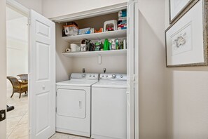 A laundry area with a washing machine and dryer, a shelf with cleaning supplies, and a wicker chair in the background. - Oceanfront Penthouse Retreat – 7th Floor at Ocean Place (Fernandina Beach)