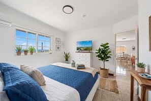 A bedroom with two beds, a large window, a painting, and a dining area visible through an open door. - Serenity at Baby Beach (Seroe Colorado)