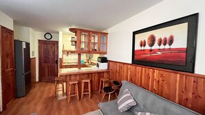 Een compacte keuken met houten kastjes, een magnetron en een koelkast. Er is een eethoek met krukken en een bank met een kussen. Een schilderij van bomen hangt aan de muur.
