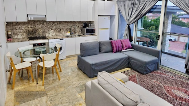 A compact living space with a kitchenette, a dining area with chairs, a sofa with cushions, and a large window with curtains. - The Bella Nest - Bella Vista (Sydney)
