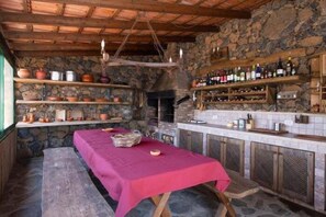 Una cocina rústica con paredes de piedra, vigas de madera, una chimenea de piedra y una larga mesa con mantel rojo.