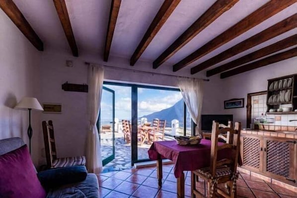 A room with a bed, a table with chairs, a view of the sea, and a kitchen area.