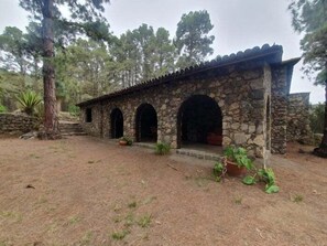 A stone building with arched doorways and a tiled roof, surrounded by trees and a dry grassy area.