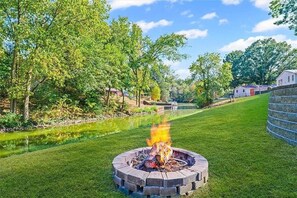 A circular stone fire pit with flames, surrounded by a well-maintained lawn and a body of water.