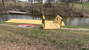Eine Holzstege, der über einen Teich führt, mit einem Haus im Hintergrund.