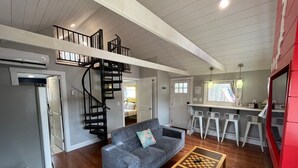 A spacious living area with a sofa, a coffee table, a spiral staircase, and a kitchen area. - Wally’s Matchbox |New|Hot Tub|Fire Pit|Close 2 All (Hawley)