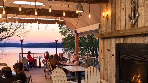 An outdoor dining area with wooden tables and chairs, a fireplace, and a view of the water. - Wally’s Matchbox |New|Hot Tub|Fire Pit|Close 2 All (Hawley)