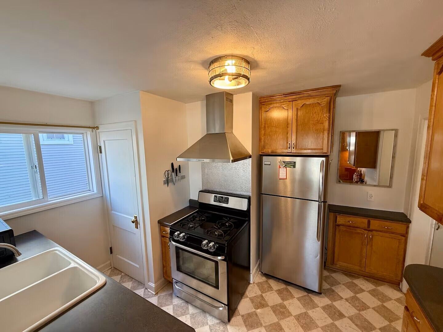A kitchen with a stove, oven, refrigerator, wooden cabinets, and a window with blinds.