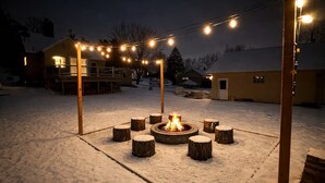A snow-covered outdoor area with a central fire pit surrounded by wooden stools, string lights overhead, and a building in the background.