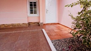Pared exterior rosa, una puerta blanca, una ventana y un pequeño jardín con guijarros y una planta.