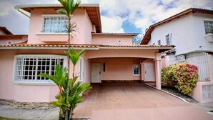 Una casa de dos pisos con techo plano, un garaje cubierto y un pequeño jardín con una palmera.