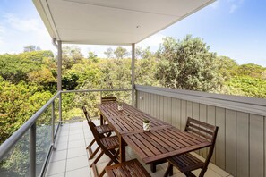 A covered balcony with a wooden table and chairs, surrounded by a railing, overlooking a lush green landscape. - Sandcastle House - Sleeps 12 steps from the beach (Point Lookout)