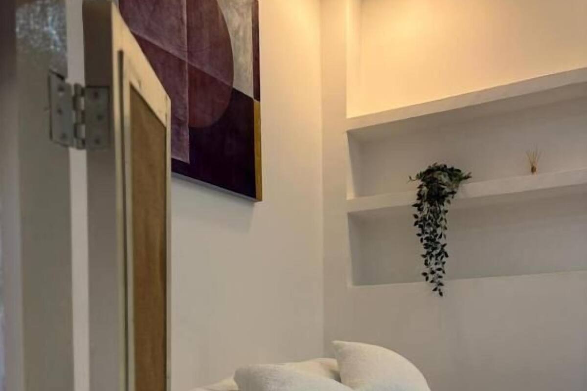 A bedroom with a bed, pillows, a wall-mounted shelf with a plant, and a painting.