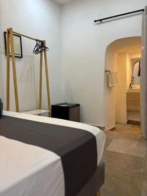 A hotel room with a bed, a wooden rack, a small desk, a trash bin, and an arched doorway leading to a bathroom. - Casa Centro Histórico Room (Cartagena)