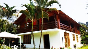 A two-story wooden house with a white exterior, surrounded by lush greenery and palm trees.