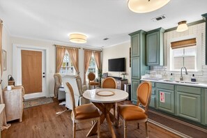 A dining area with a round table and chairs, a kitchen with green cabinets, and a television on the wall. - Tiny Tide Cottage· Golf Cart Add · Stroll Downtown (Ocean Springs)