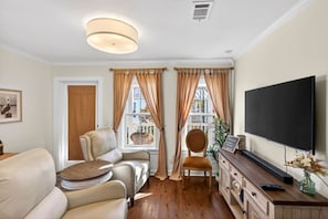 A living room with a sofa, armchairs, a television, and a large window with curtains. - Tiny Tide Cottage· Golf Cart Add · Stroll Downtown (Ocean Springs)
