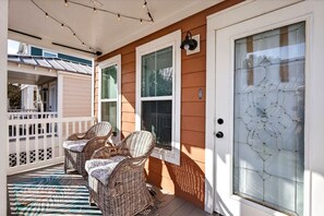 A porch with wicker chairs and a patterned rug, a white door with a decorative glass panel, and string lights above. - Tiny Tide Cottage· Golf Cart Add · Stroll Downtown (Ocean Springs)
