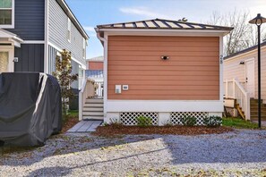 A single-story mobile home with a covered grill, a small porch, and a gravel driveway. - Tiny Tide Cottage· Golf Cart Add · Stroll Downtown (Ocean Springs)