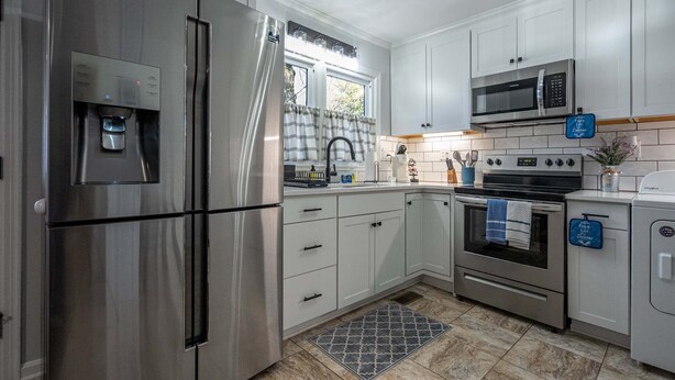 A modern kitchen with stainless steel appliances, white cabinetry, a tiled backsplash, and a window with curtains.