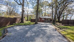 A gravel driveway leading to a small building with a porch, surrounded by trees and a wooden fence.