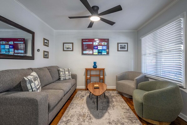 A living room with a gray sofa, a green armchair, a wooden coffee table, a ceiling fan, a TV mounted on the wall, and a window with blinds.