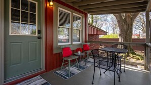 A covered porch with a green door, two windows with blinds, a small table with chairs, and a tree in the background.