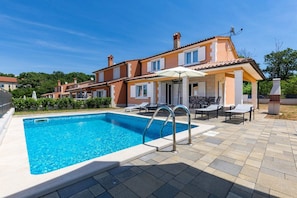 Ein Schwimmbecken mit klarem, blauem Wasser, umgeben von einer Terrasse mit Liegestühlen und einem Grill.
