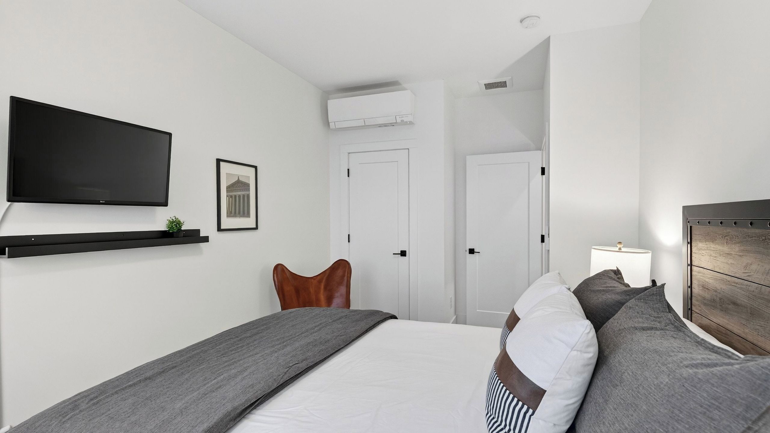 A modern bedroom with a bed, a mounted TV, a wall-mounted air conditioner, and a door leading to another room.