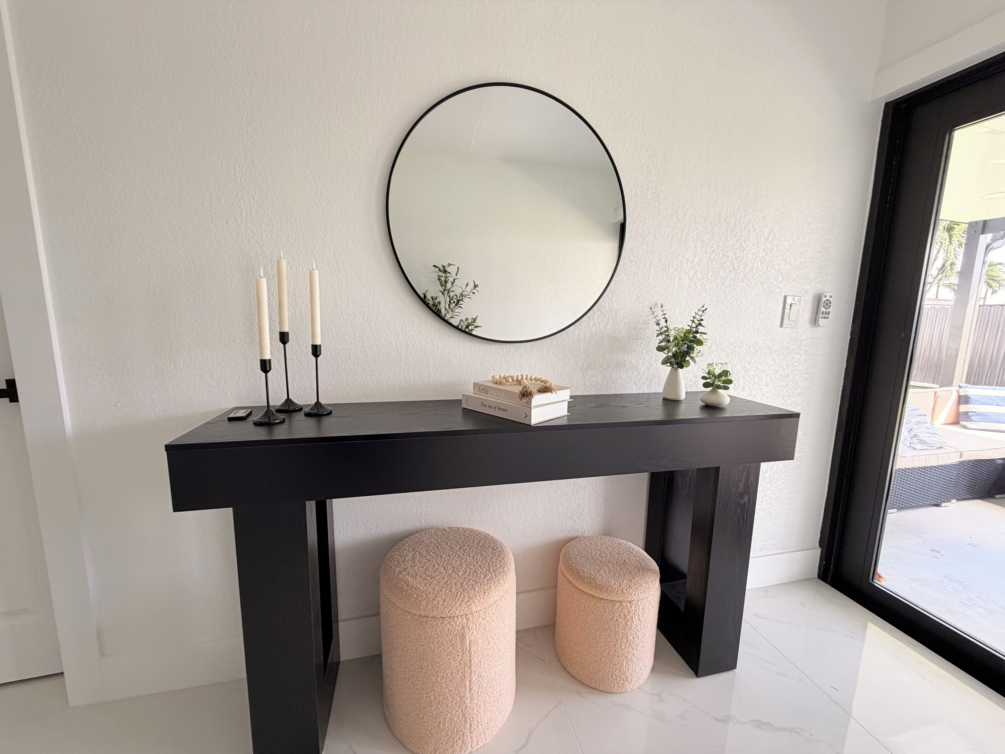 A modern interior with a black console table, a round mirror, two candles, a small plant, and two ottomans.