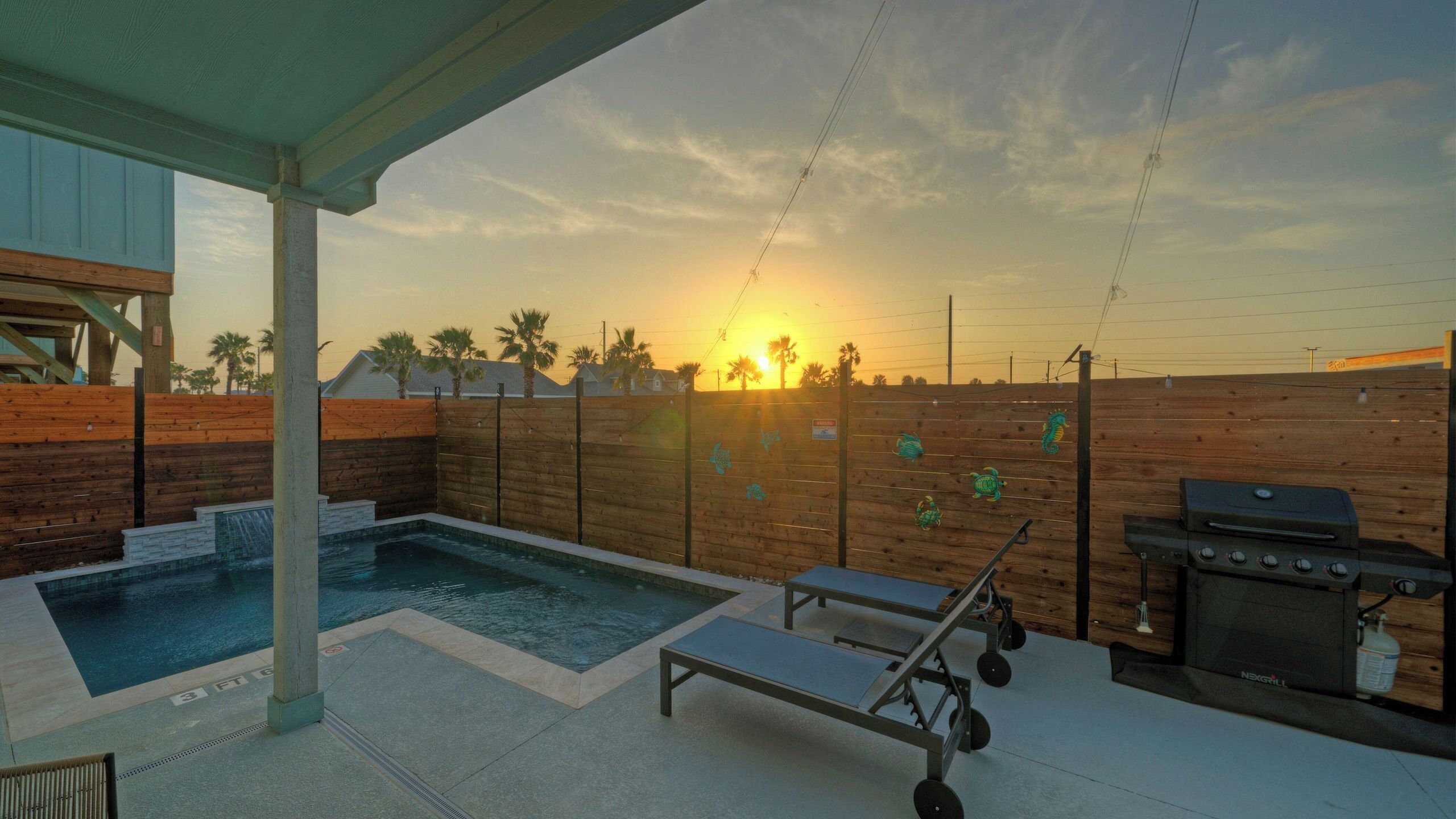A backyard with a swimming pool, lounge chairs, a grill, and a wooden fence.
