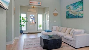 A living room with a white sofa, a black coffee table, a large abstract painting, and a door labeled 'PARADISE.' - Mermaid's Oasis by Avantstay Spacious Port A Home + Private Pool + Firepit + Games Near the Beach (Port Aransas)