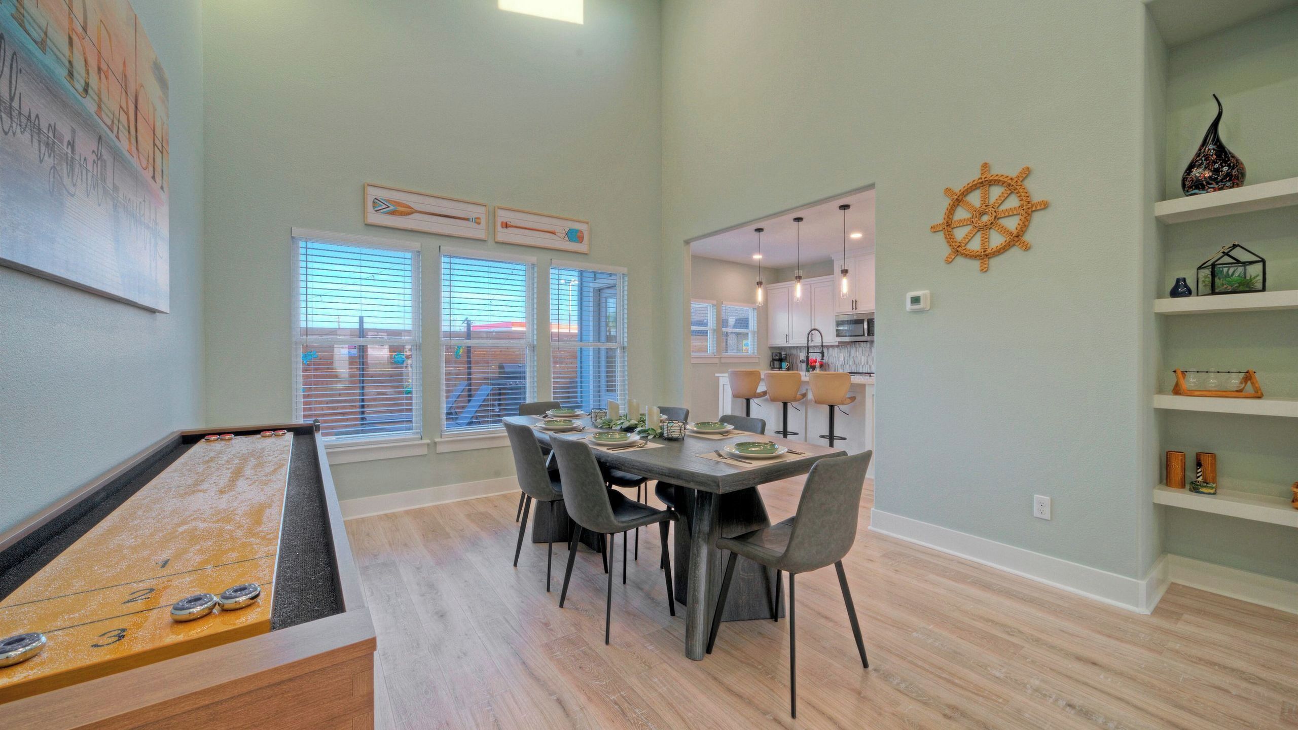 A dining area with a pool table, a kitchen in the background, and a view through the windows.