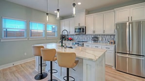 A modern kitchen with white cabinets, stainless steel appliances, and a central island with bar stools. - Mermaid's Oasis by Avantstay Spacious Port A Home + Private Pool + Firepit + Games Near the Beach (Port Aransas)