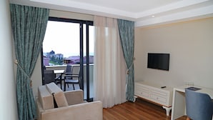 A hotel room with a sofa, a dining table with a plate of food, a desk with a chair, a TV, and a view through a large window.
