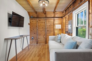 Une pièce chaleureuse avec un plafond en bois, un téléviseur à écran plat fixé au mur, un canapé avec des coussins et une fenêtre donnant sur un paysage verdoyant.