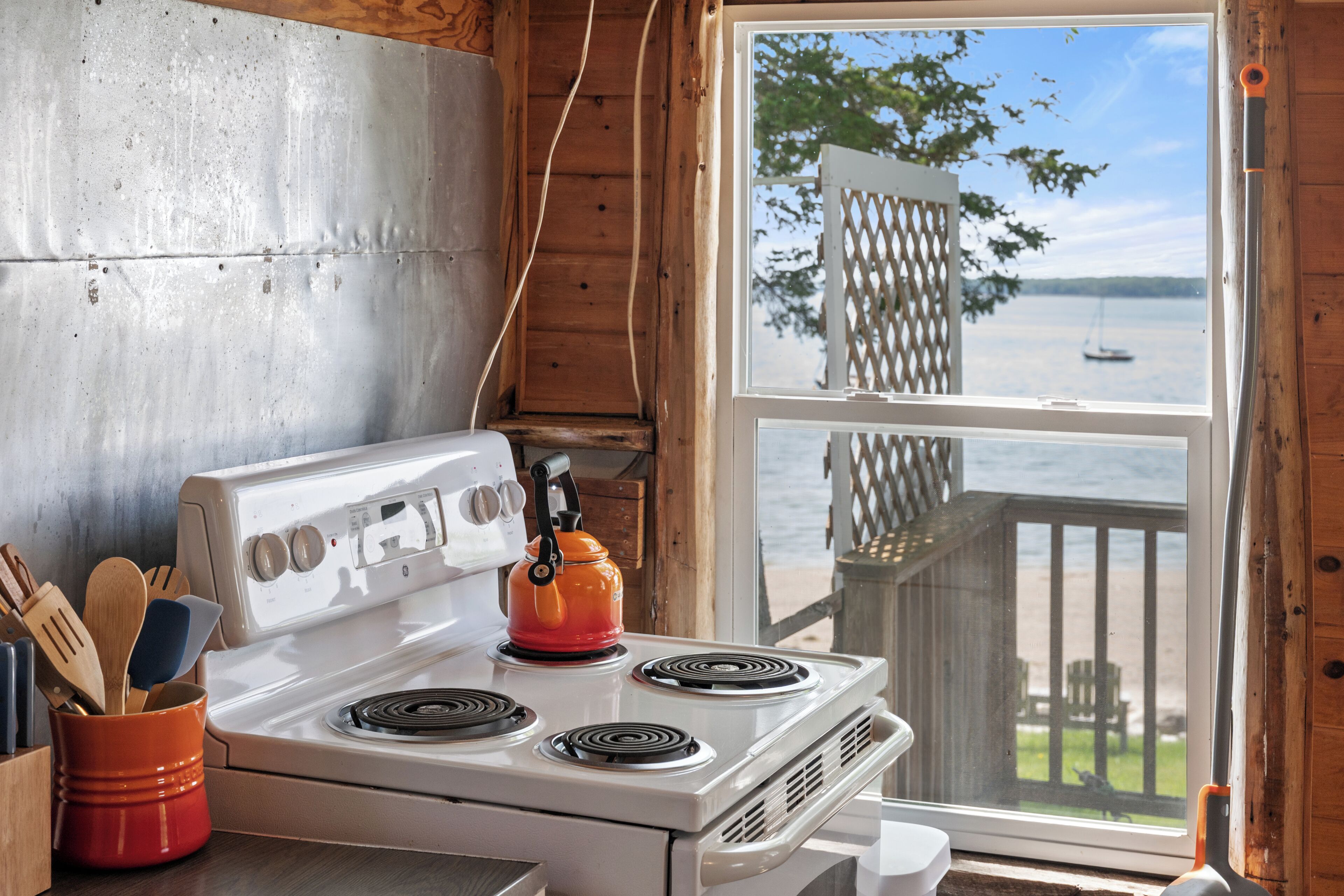 A kitchen with a stove, a kettle, and a window overlooking a body of water.