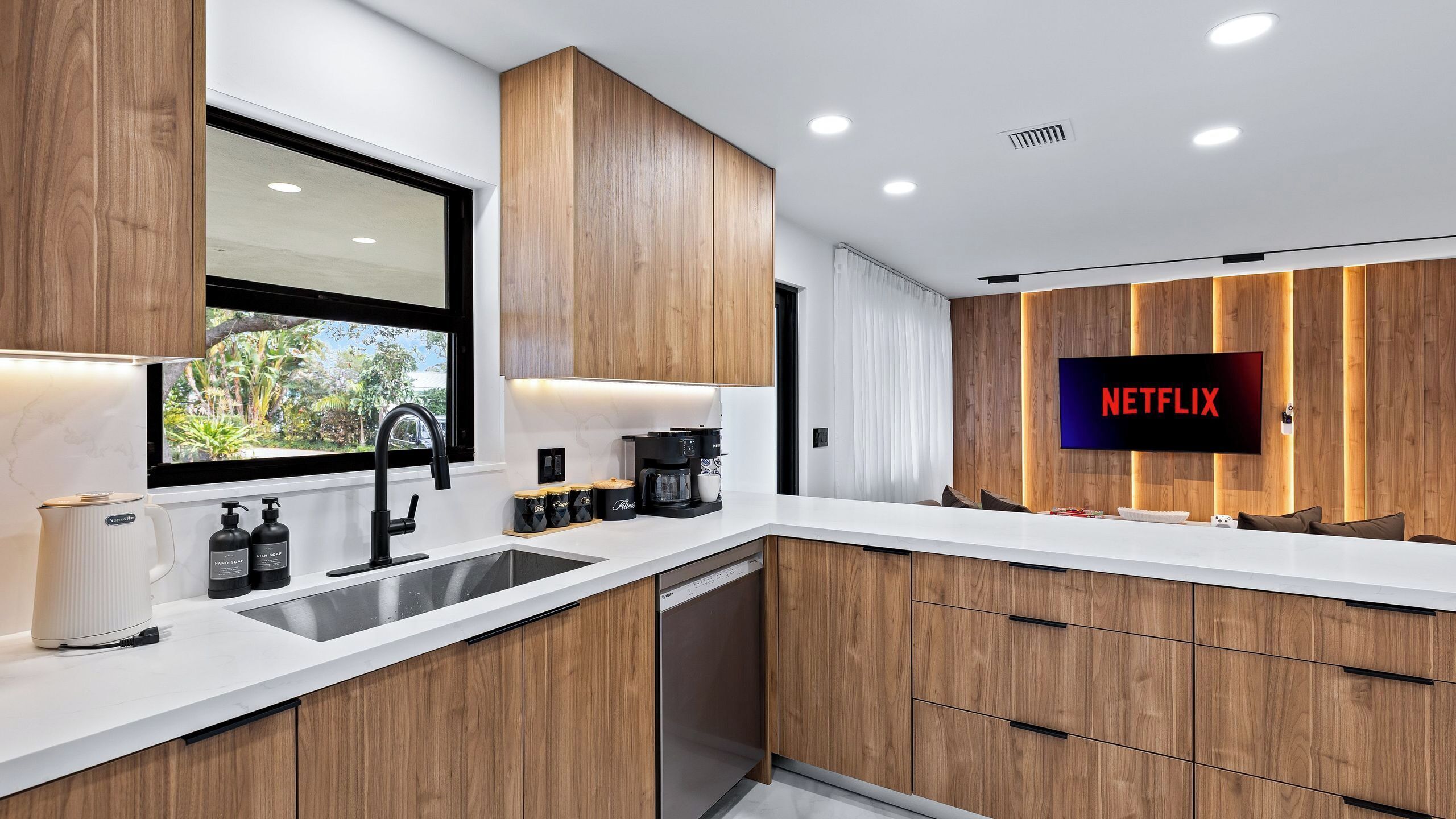 A modern kitchen with wooden cabinets, a built-in dishwasher, a coffee maker, and a large flat-screen TV displaying 'NETFLIX'.