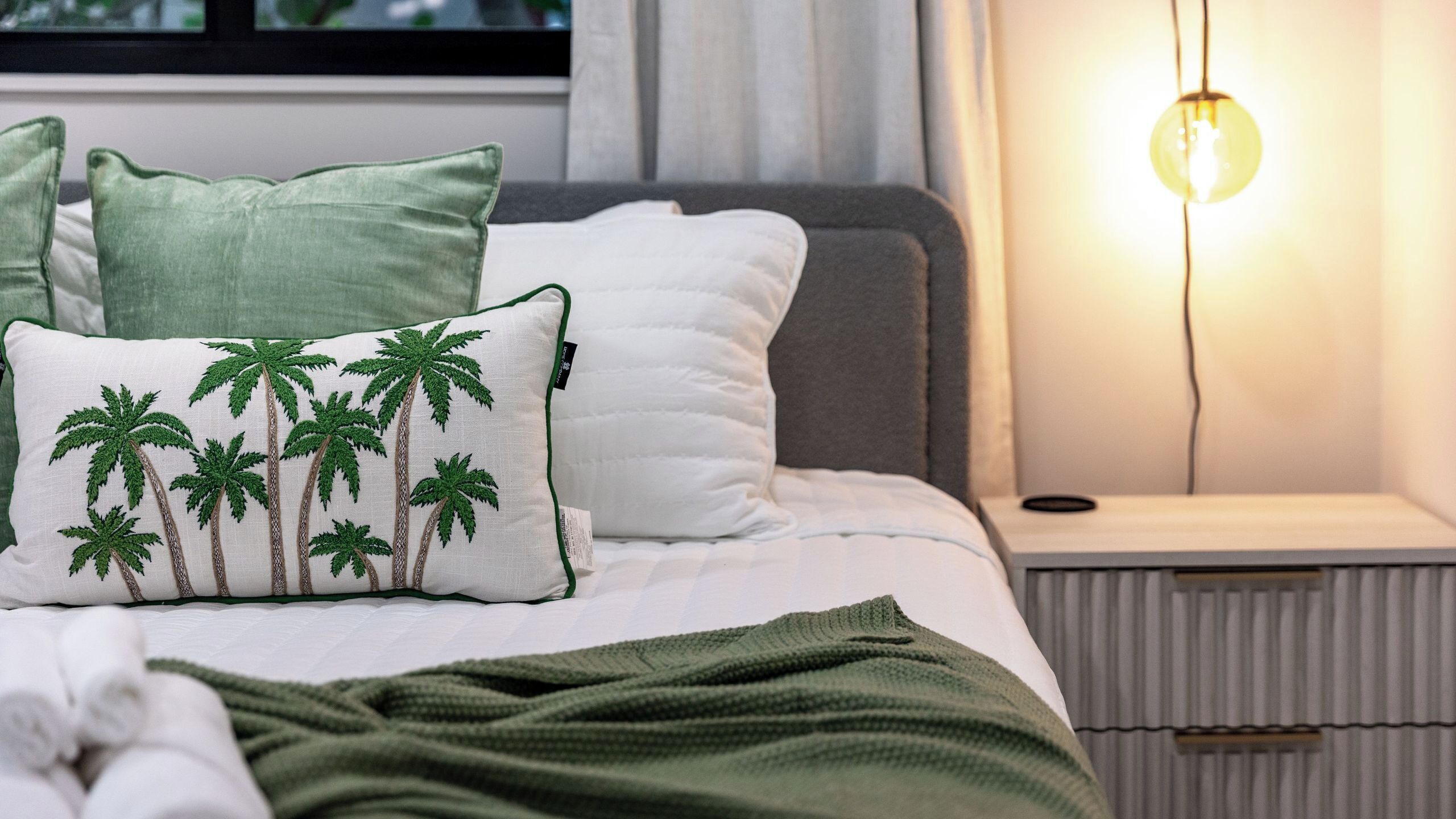 A neatly made bed with a green blanket, a pillow with palm tree embroidery, and a bedside lamp.