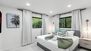 A modern bedroom with a bed, curtains, a wall-mounted light, and a window with a view of greenery. - Soleil Oasis by Avantstay Screened Pool + Outdoor Lounge + Firepit Near the Beach (Fort Lauderdale)