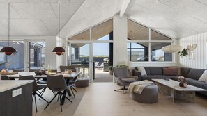 Amplia sala de estar con una mesa de comedor, sillas, un sofá y una mesa de centro. Grandes ventanales ofrecen luz natural y vistas al exterior.