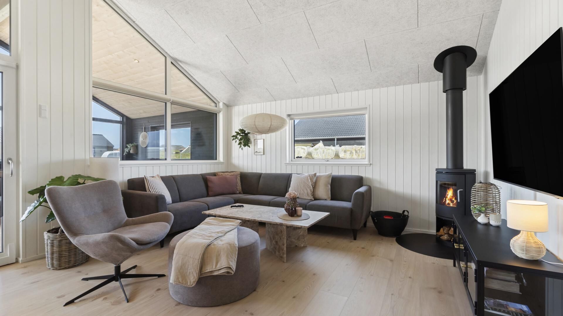 Un salon moderne avec un canapé d’angle, un pouf rond, une cheminée et une grande fenêtre.