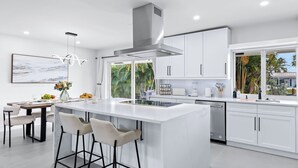 A modern kitchen with white cabinetry, a central island with bar stools, a large window with a view of palm trees, and a painting on the wall. - Soleil Breeze by Avantstay Pool + Canalfront Home + Outdoor Dining Canal Deck + Kayak 10 Mins to the Beach (Fort Lauderdale)