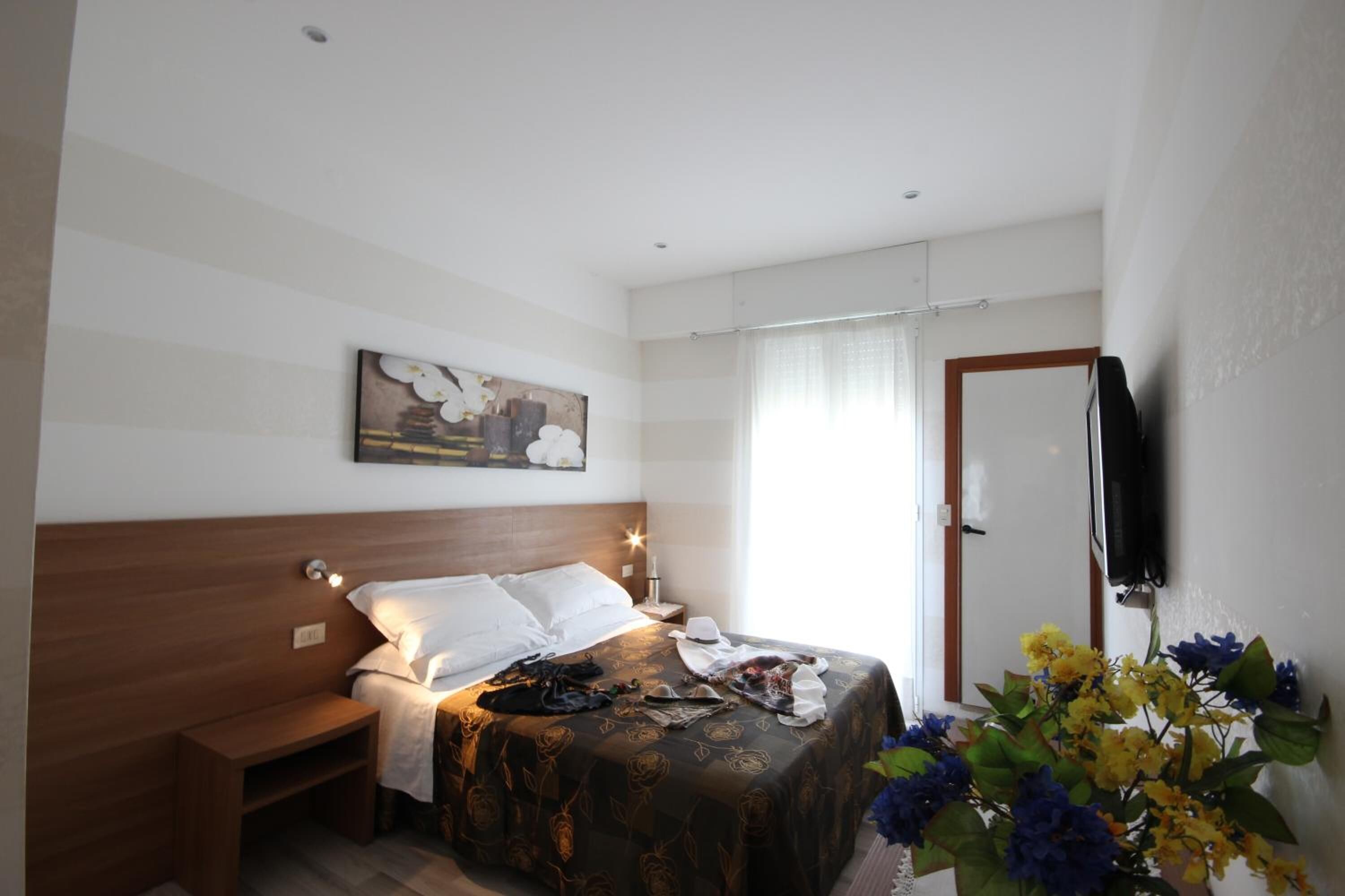 A bedroom with a bed, bedside table, nightstand, wall-mounted TV, and a window with curtains.