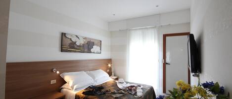 A bedroom with a bed, bedside table, nightstand, wall-mounted TV, and a window with curtains.