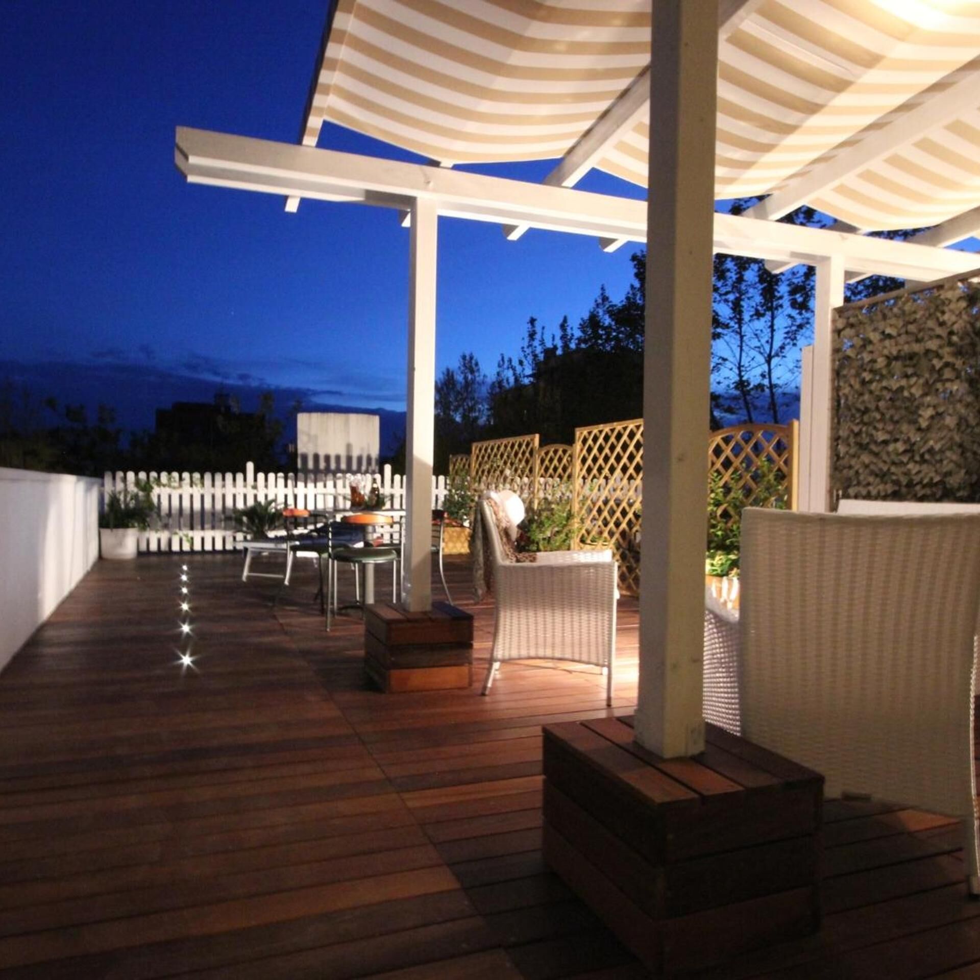 A covered outdoor patio with wooden flooring, white wicker furniture, and a white fence.