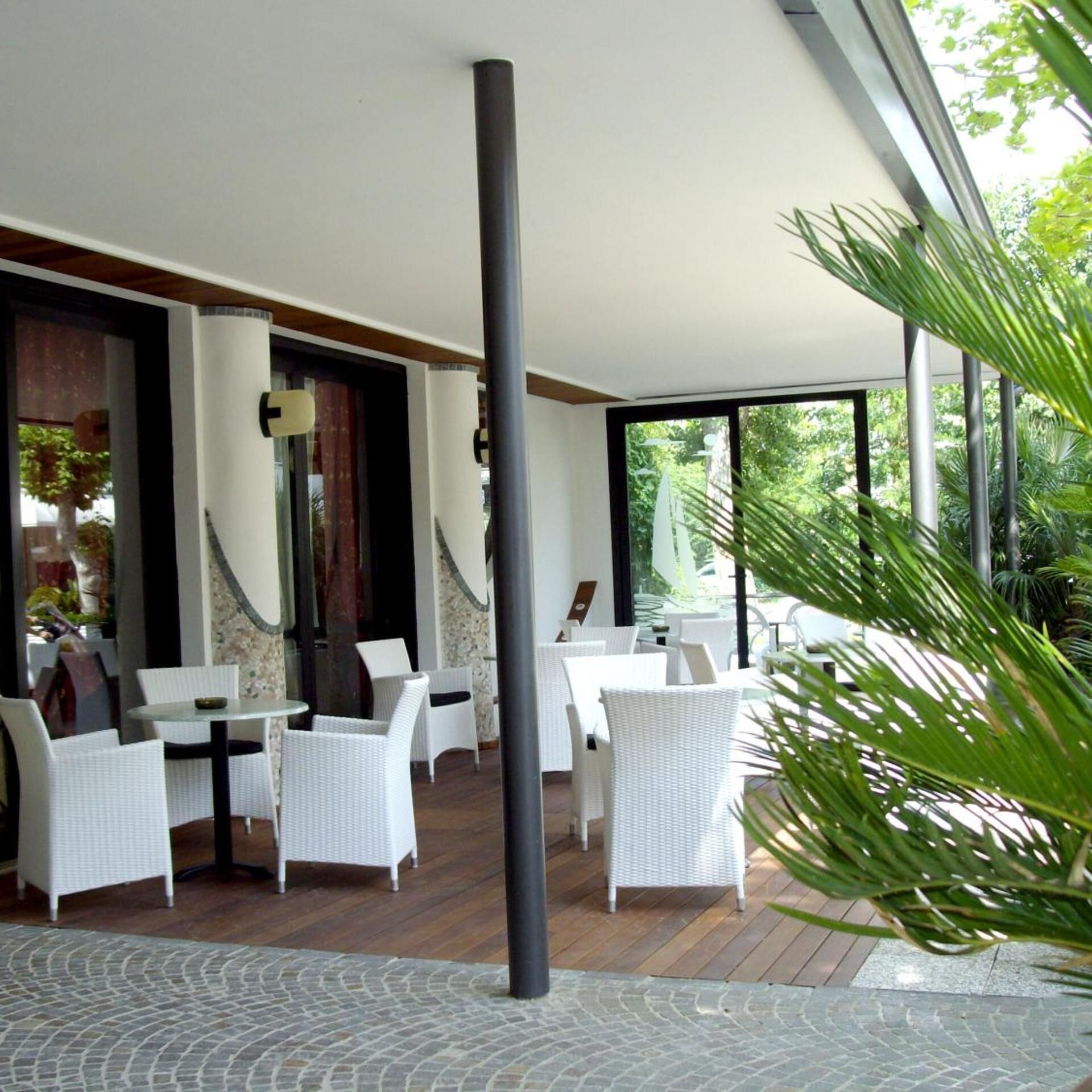 An outdoor patio with white wicker furniture, a large black pole, and a view of greenery through large windows.