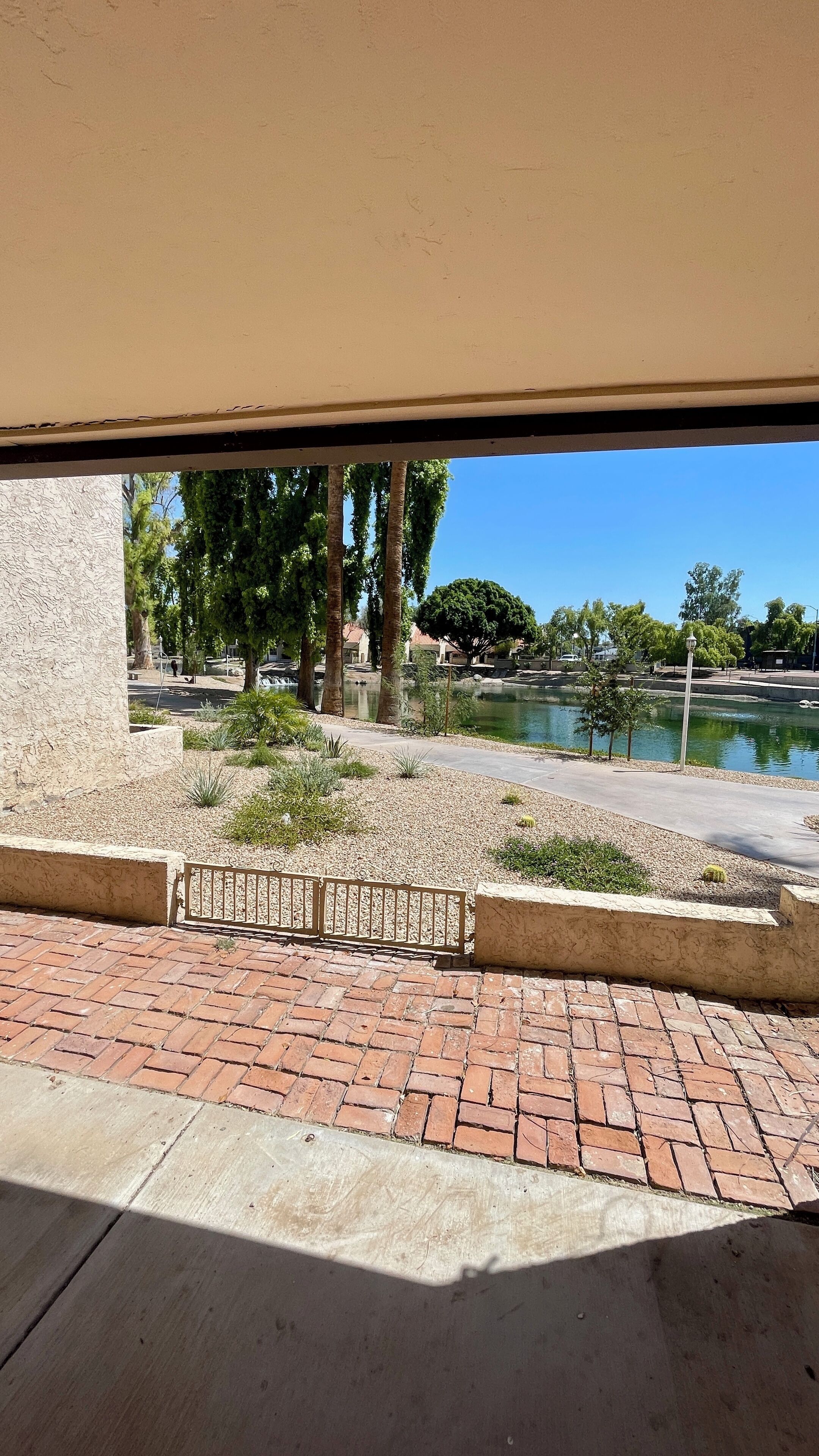A paved walkway with a brick pattern, a metal fence, and a view of a body of water with trees and a clear sky.