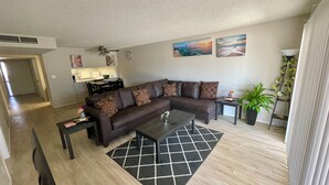 Un salon comprenant un canapé d’angle, une table basse, un tapis et un coin repas avec une cuisine en arrière-plan.