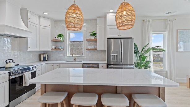 Una cocina moderna con armarios blancos, una isla central con taburetes, un refrigerador de acero inoxidable y lámparas colgantes de mimbre.